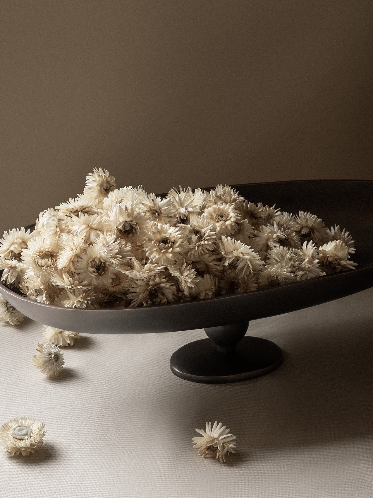 A close-up shot of the Pavot Centerpiece, a dark pedestal bowl, overflowing with a mound of dried, off-white flowers.