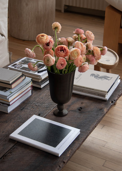 The Audo Pavot Vase, a dark chalice-shaped vessel, holds pink ranunculus flowers on a rustic wooden coffee table with books.