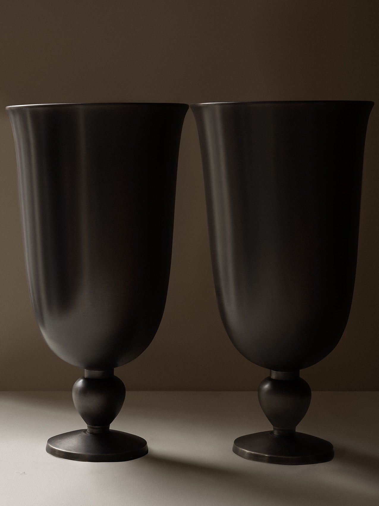 A close-up of two Pavot Vases by Audo, featuring a dark, matte bronzed finish and a classic goblet shape with a flared rim.