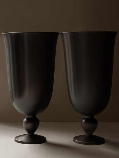 A close-up of two Pavot Vases by Audo, featuring a dark, matte bronzed finish and a classic goblet shape with a flared rim.