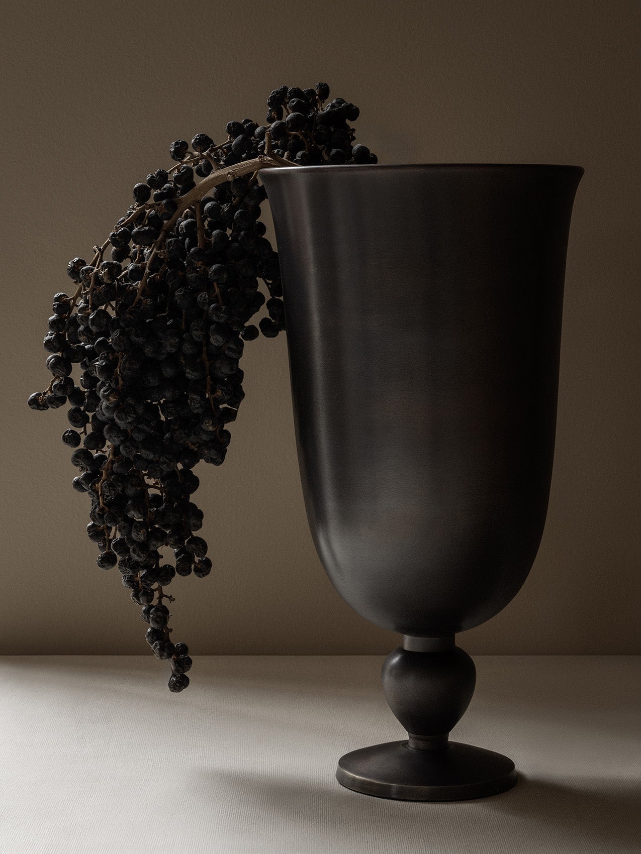 The Pavot Vase, a chalice-shaped vase in a dark metallic finish, styled with a bunch of dried berries draped over the edge.
