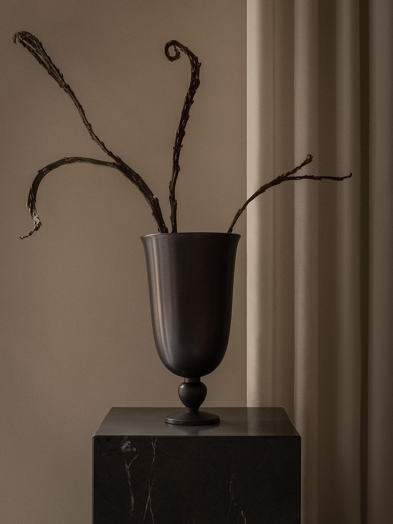 The black, chalice-shaped Pavot Vase holding sculptural, dried branches, on a dark marble plinth against a neutral backdrop.