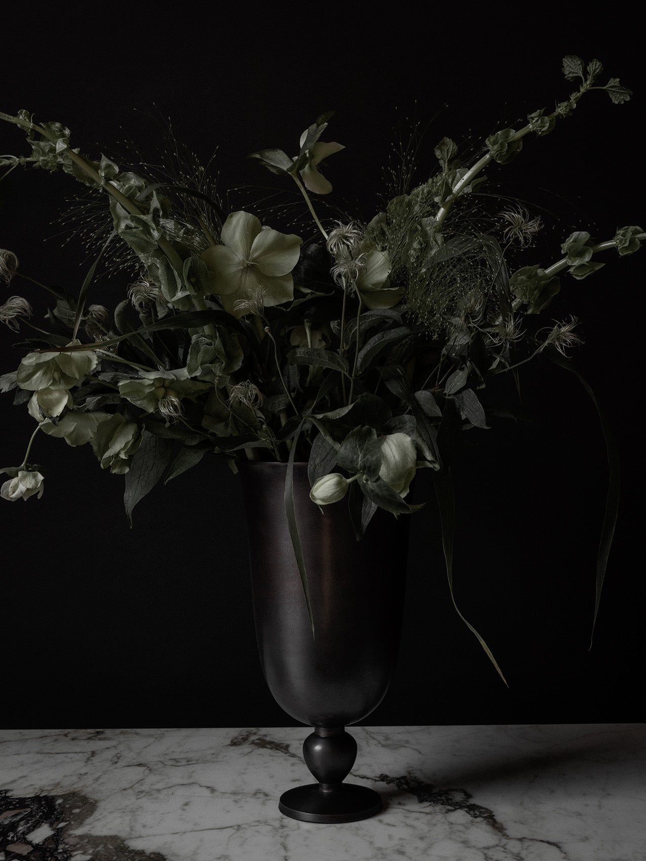 The dark metal Pavot Vase styled with a large bouquet of pale green flowers and foliage on a marble tabletop.