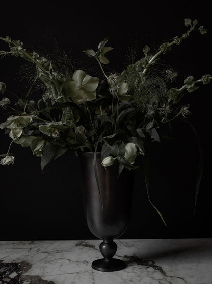 The dark metal Pavot Vase styled with a large bouquet of pale green flowers and foliage on a marble tabletop.