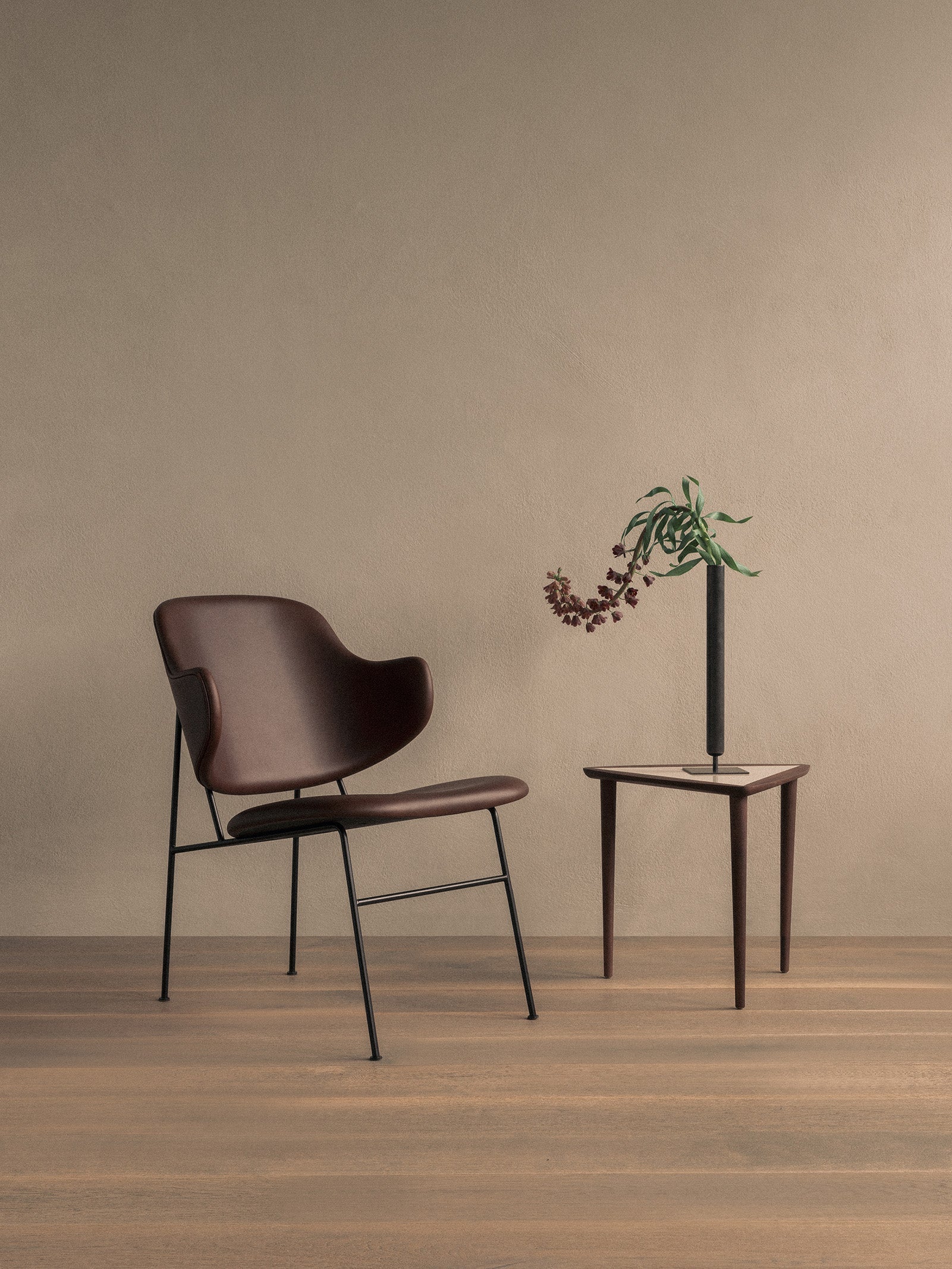 The brown upholstered Penguin Lounge Chair is next to a wooden Umanoff side table with a black Stance Vase and flowers.
