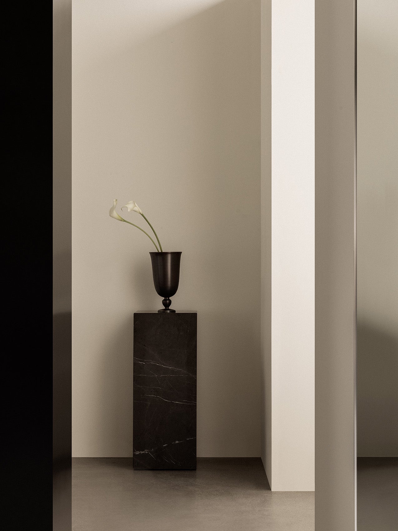 A tall Plinth Pedestal in black marble stands in a minimalist setting. The dark Pavot Vase holds two white calla lilies.