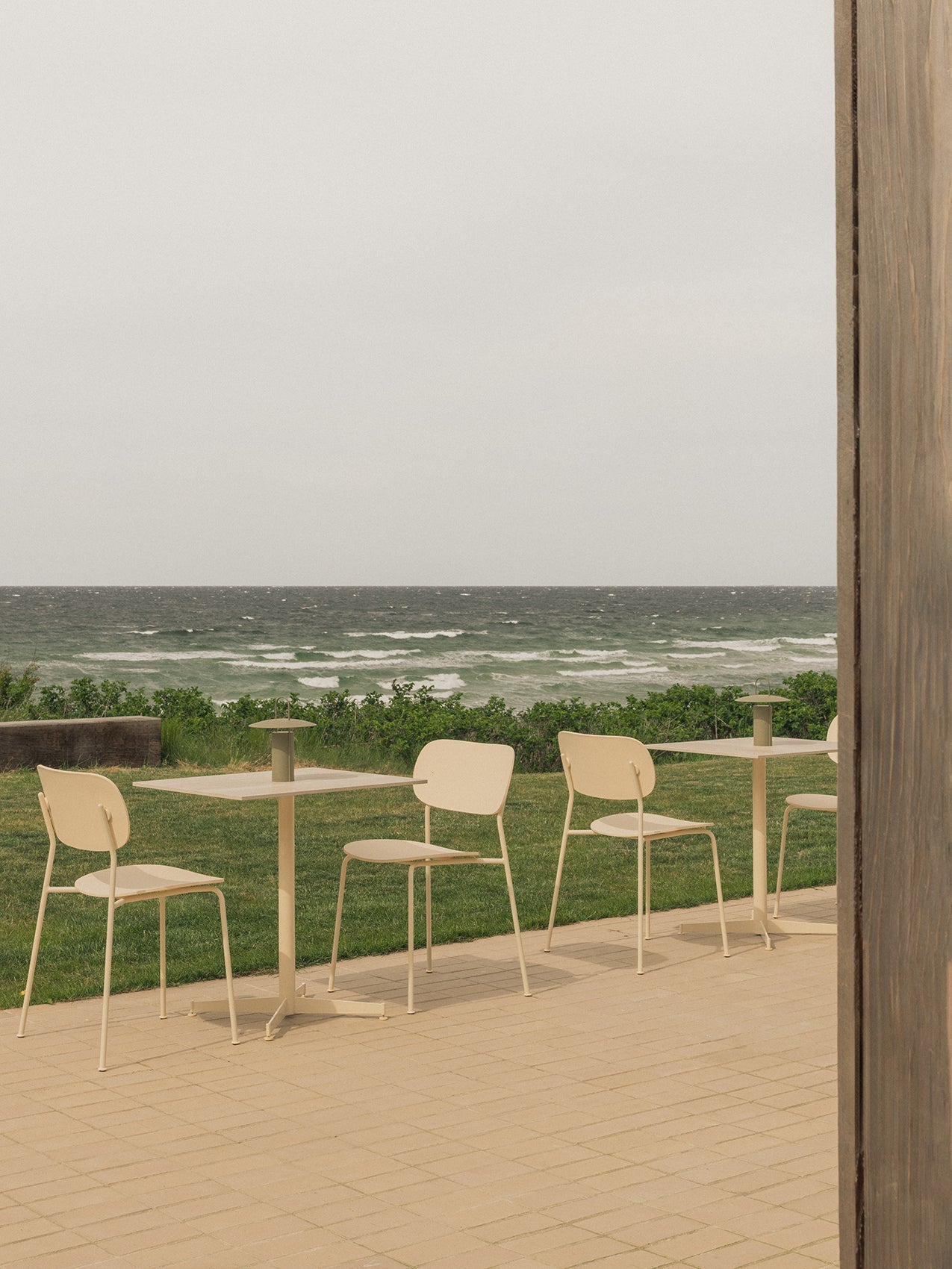 An outdoor patio with ivory Nook Dining Tables, Co Dining Chairs, and an olive Ray Table Lamp, set against a windy sea.