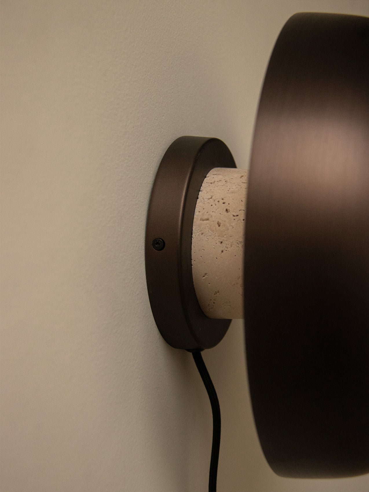 A close-up side view of the Reverse Wall Lamp, detailing its bronzed metal fixture and porous travertine stone base.