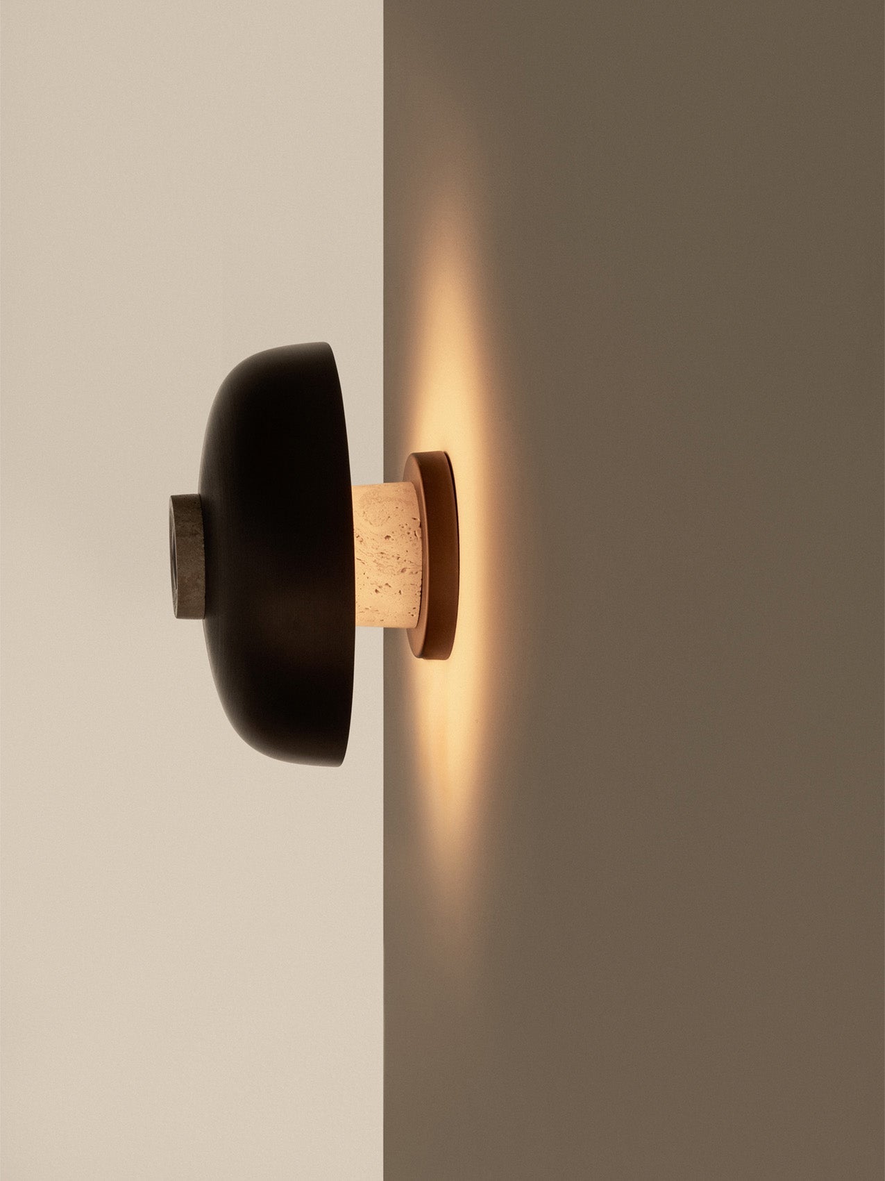 The Reverse Wall Lamp, Dimmer on Lamp, at a corner, casting a warm glow from its travertine base and bronzed aluminium shade.