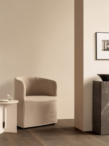 In a beige room, a Tearoom Club Chair with a loose cover sits by an Androgyne Side Table and a tall, dark marble Plinth.