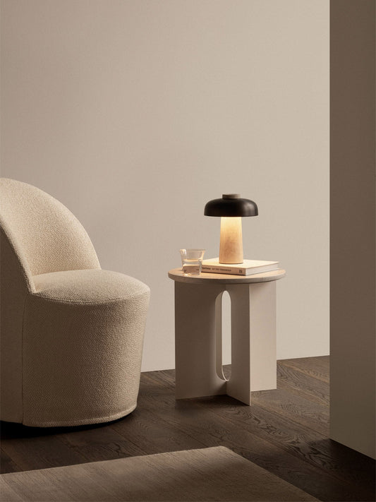 A cream Tearoom Club Chair with Swivel sits beside an Androgyne Side Table holding a lit Reverse Table Lamp Portable.