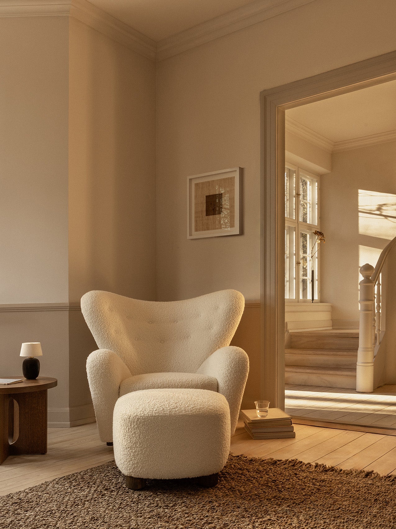A sunlit room with The Tired Man Lounge Chair and Ottoman in white fabric, beside a wooden side table and a small table lamp.