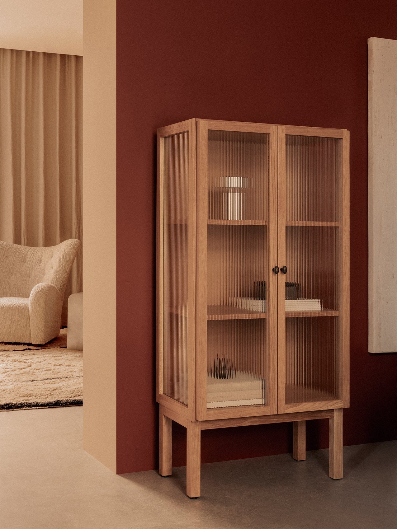 Audo's oak Curiosity Cabinet with reeded glass doors against a red wall, with The Tired Man armchair in the background.