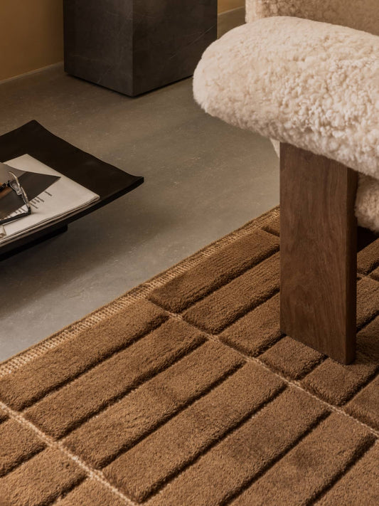 A close-up of a brown Tile Rug by a sheepskin Brasilia Lounge Chair with a Releve Platter and dark marble Plinth Tall.