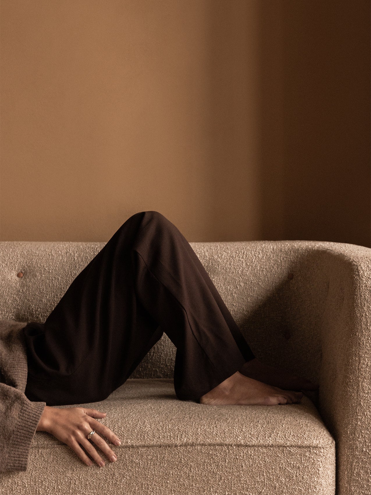 A person in dark trousers and a brown jumper relaxing on the light-coloured, textured Vilhelm Sofa against a warm brown wall.