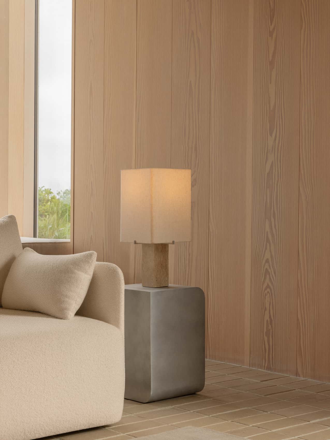 The lit Nonna Table Lamp on a grey Volume Side Table in a corner with wood-panelled walls and a cream sofa.