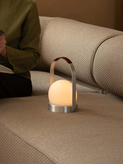 A glowing Carrie Portable Lamp with a brushed aluminium frame and leather handle rests on a beige textured sofa.