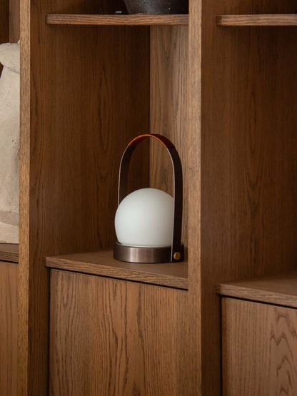Carrie Portable Lamp with a leather handle and opal glass shade, resting inside a warm wooden shelving unit.