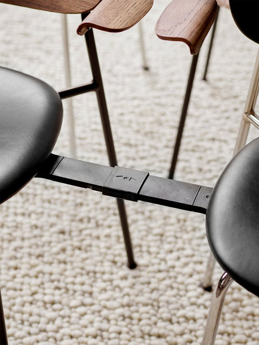 A close-up of the black metal Co Chair Row Connector linking two chairs with black seats on a light-coloured textured rug.