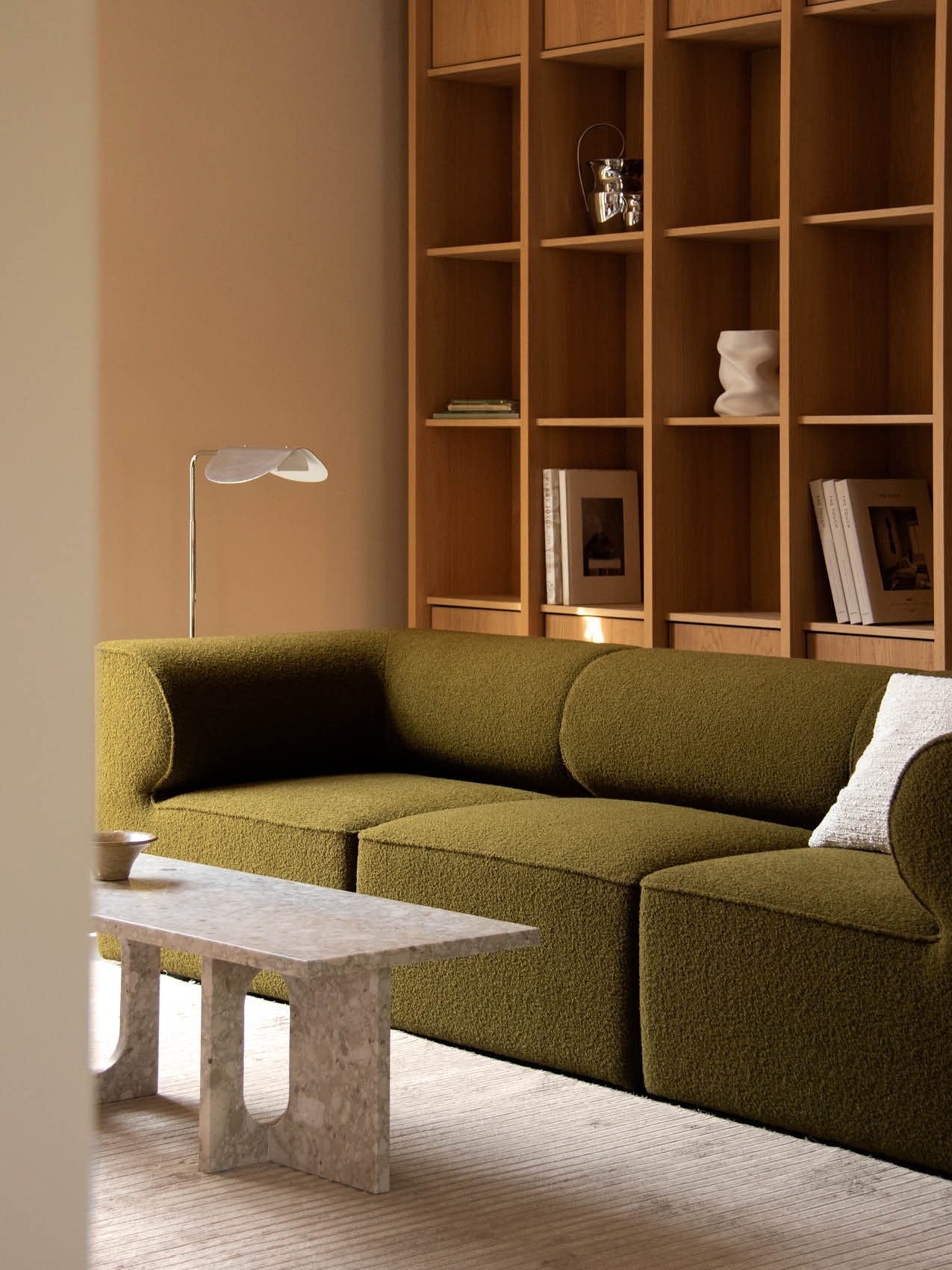 The olive green Eave Modular Sofa in a living room with a marble coffee table, a silver floor lamp, and a wooden bookcase.