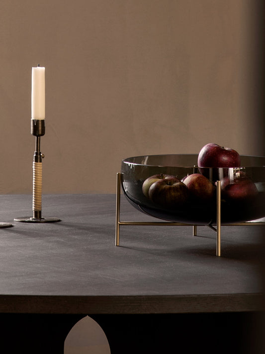 Echasse Bowl, a smoked glass vessel on a brass stand, filled with red apples and placed on a dark tabletop beside a candle.