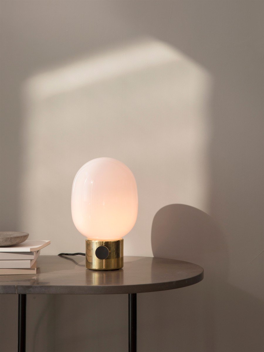 The JWDA Table Lamp in polished brass with an illuminated opal glass shade, on a dark grey table next to a stack of books.