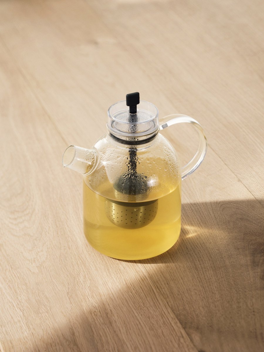 A clear glass Kettle Teapot filled with pale yellow tea, with a metal infuser inside, sitting on a light wood surface.
