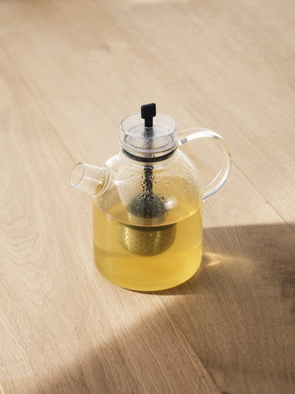 A clear glass Kettle Teapot filled with pale yellow tea, with a metal infuser inside, sitting on a light wood surface.