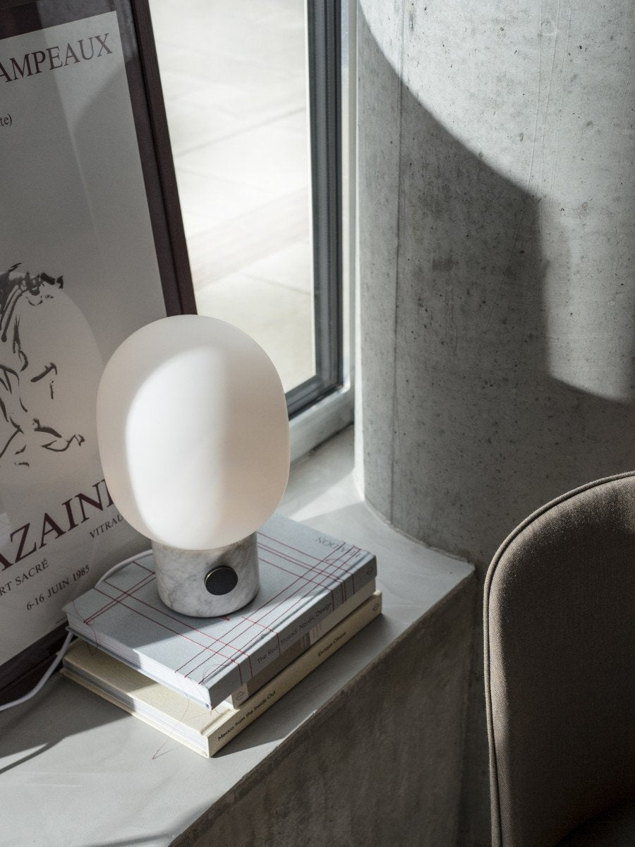 The JWDA Table Lamp, with a marble base and opal glass shade, styled on a stack of books on a sunlit concrete windowsill.