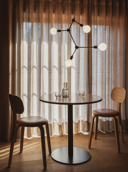A round Harbour Column Dining Table set with two glasses and a carafe, placed between two wooden chairs under a chandelier.