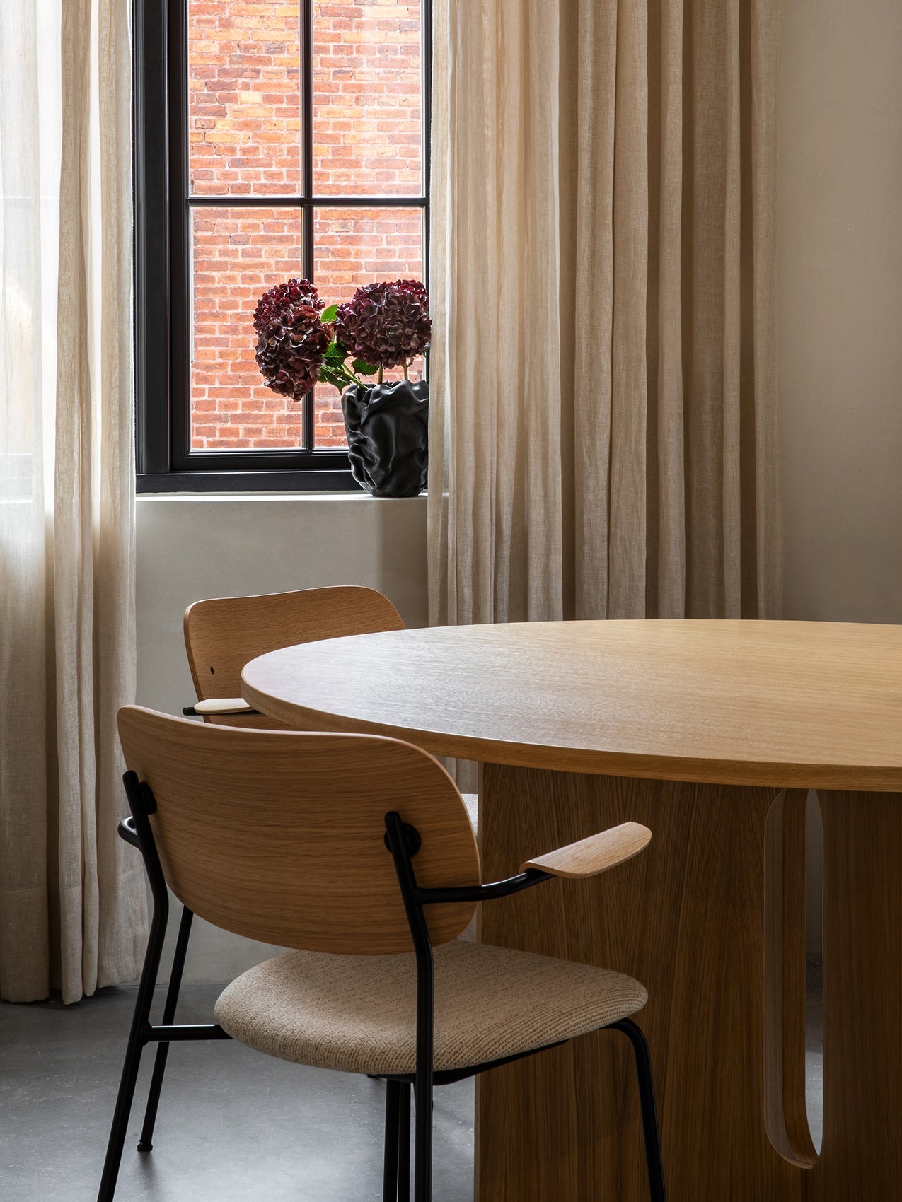 Two Co Dining Chairs with light wood backs and armrests tucked into a large Androgyne Dining Table in front of a window.