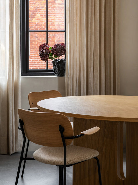 Two Co Dining Chairs with light wood backs and armrests tucked into a large Androgyne Dining Table in front of a window.