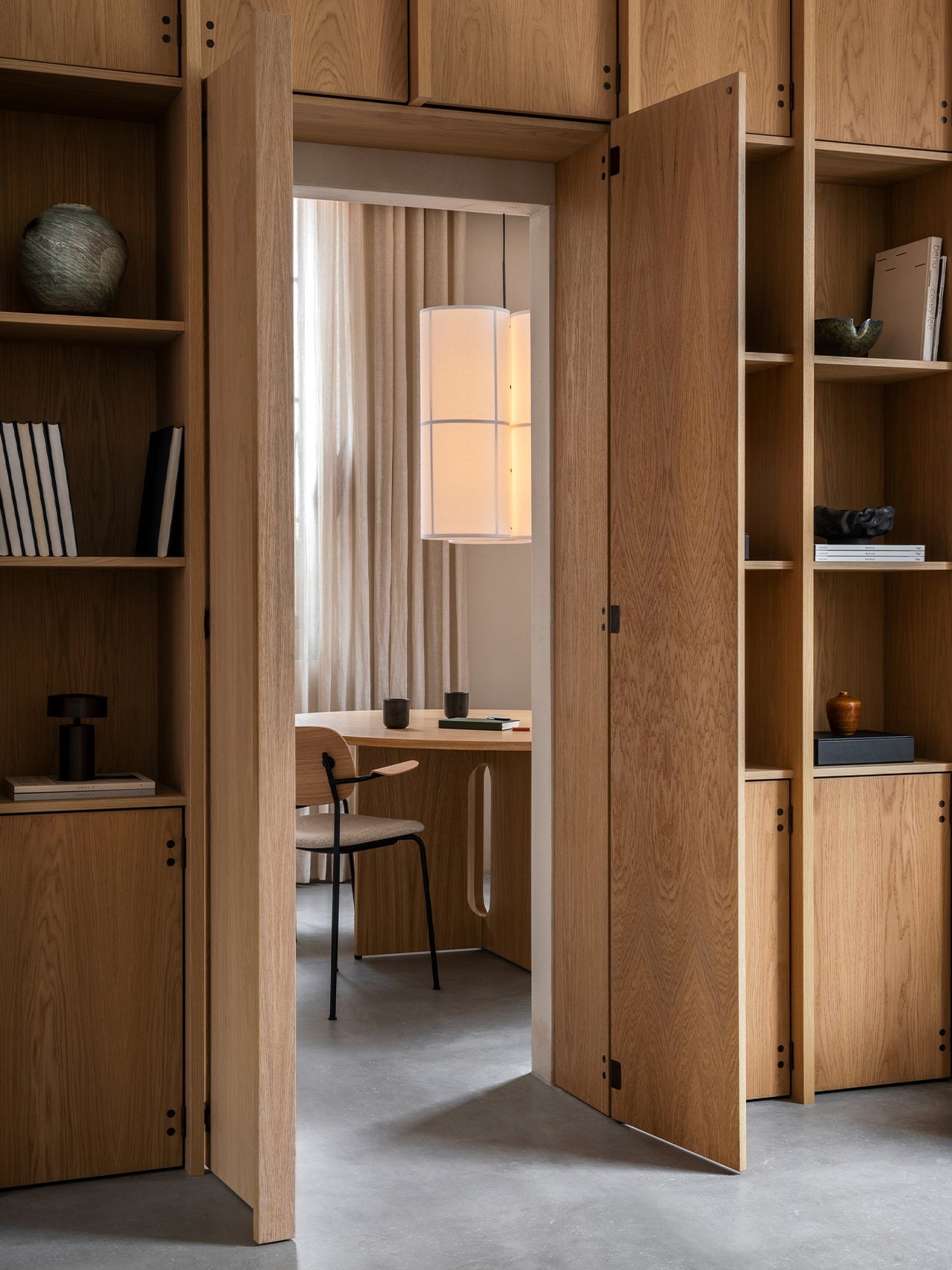 View through an open wooden bookcase into a dining room with the Androgyne Dining Table, Co Chair, and Hashira Pendant lamp.