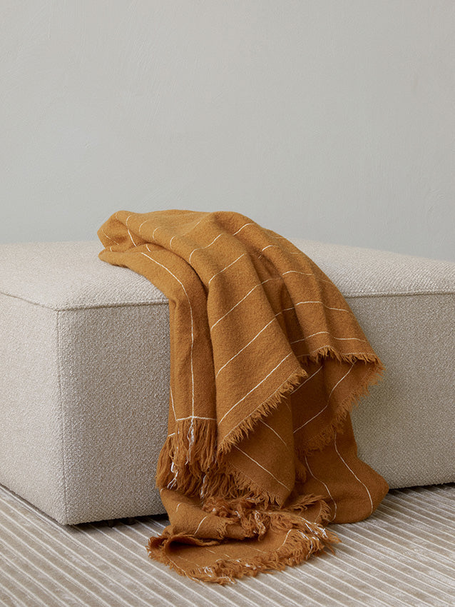 A mustard-yellow Battus Throw with frayed edges and white stripes, casually draped over a light-coloured Eave Modular Pouf.