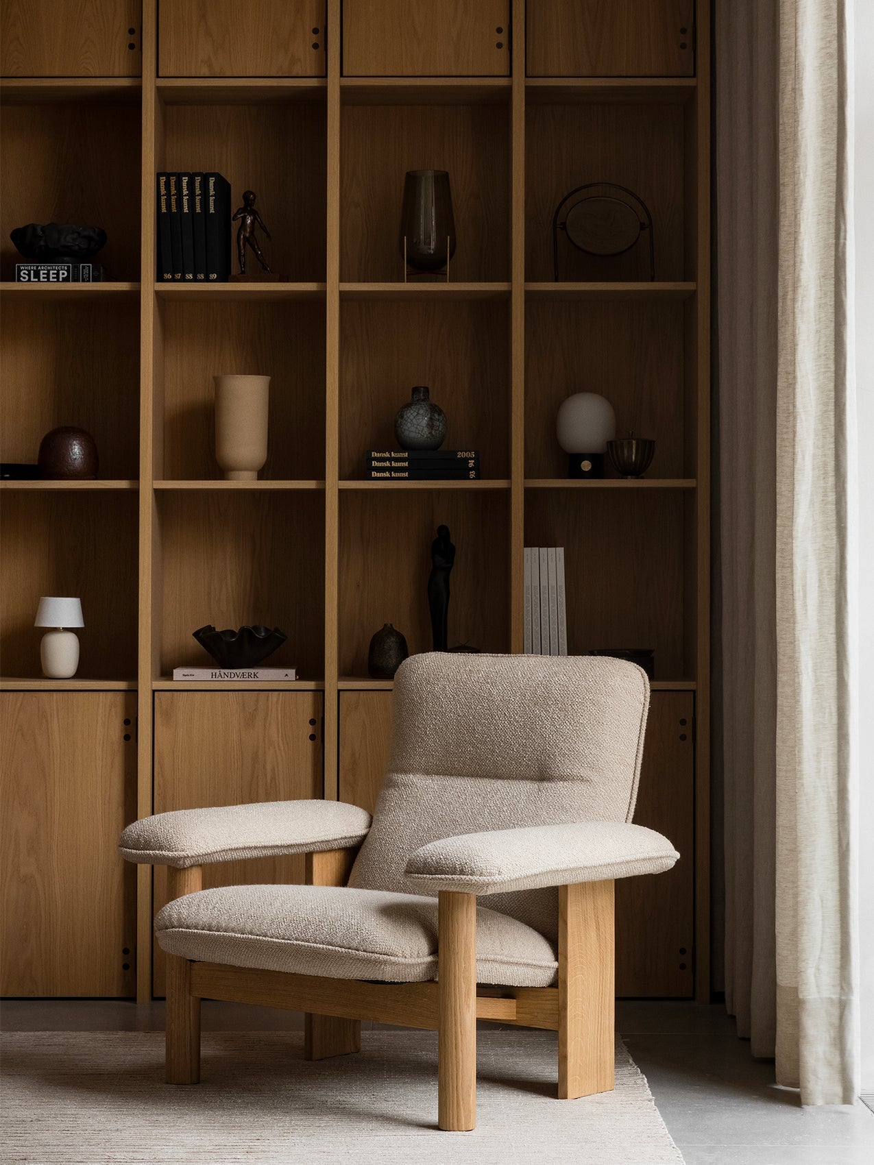 A Brasilia Lounge Chair in light bouclé fabric and oak frame sits on a Houkime Rug before a bookcase with a Torso Lamp.