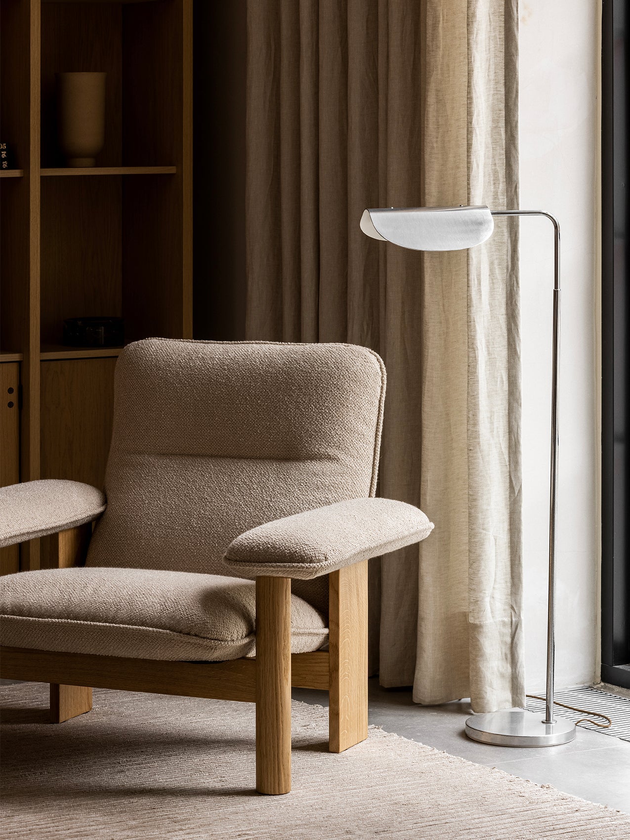 A close-up of the Brasilia Lounge Chair in beige upholstery and an oak frame, placed on a Houkime Rug next to a floor lamp.