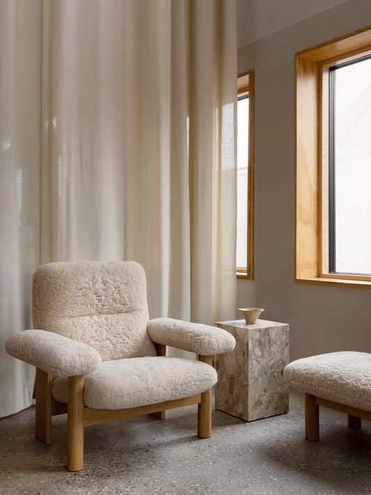 The Brasilia Lounge Chair and Ottoman in sheepskin sit by a Plint Cubic marble table in a serene room with large windows.