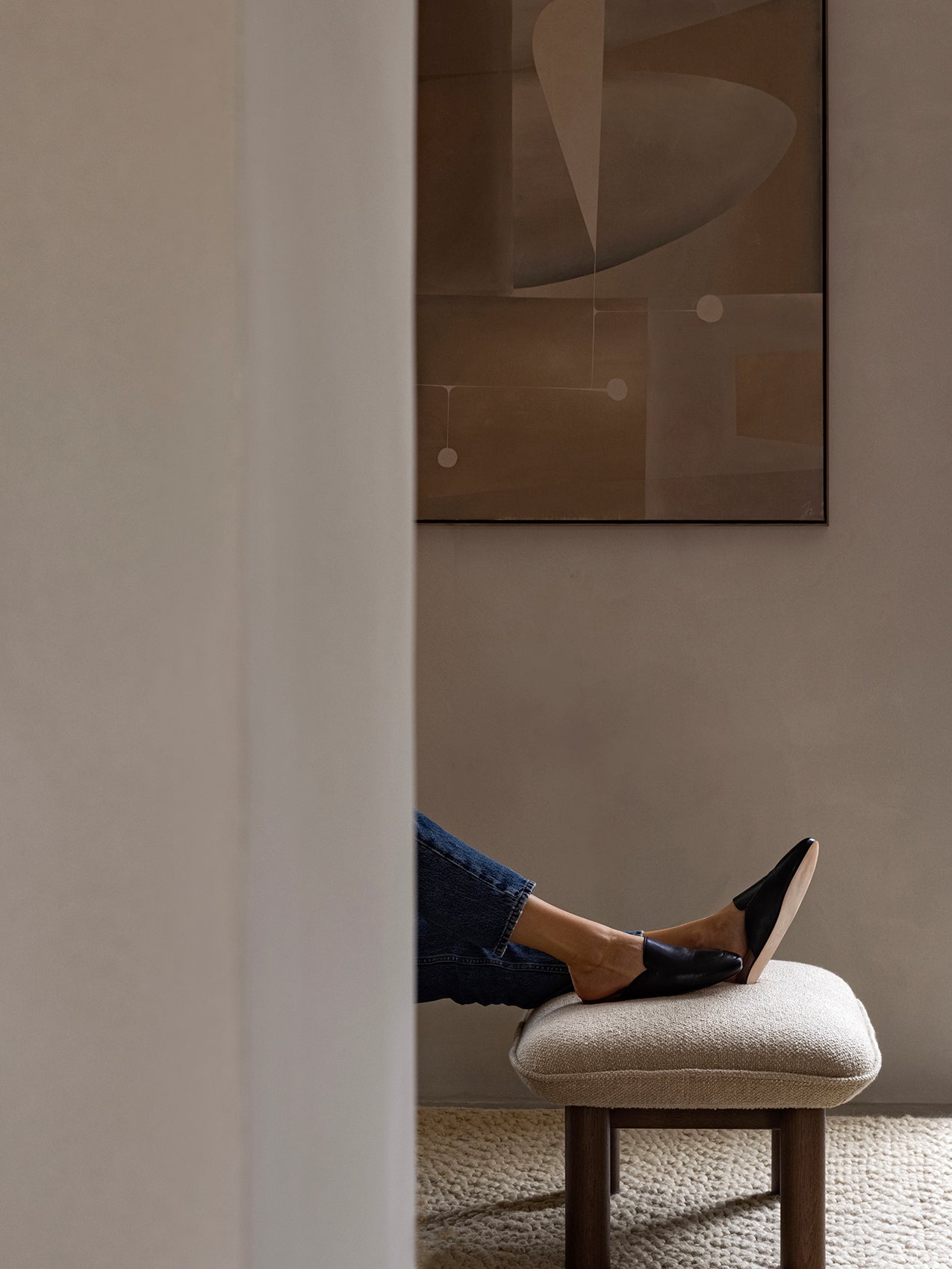 A person resting their feet, clad in black slippers, on the light-coloured, textured Brasilia Ottoman with dark wooden legs.