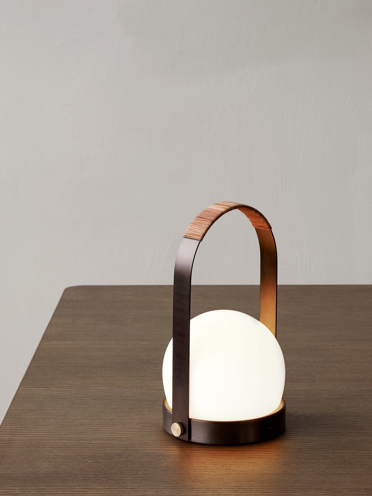 A close-up of the Carrie Table Lamp in bronzed brass, its leather-wrapped handle and lit opal glass shade on a wooden table.