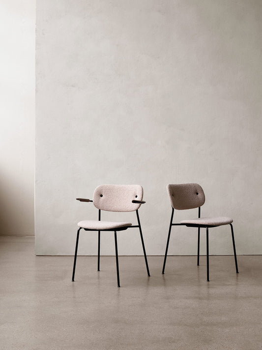 Two fully upholstered Co Dining Chairs with black legs stand on a concrete floor against a textured, off-white wall.