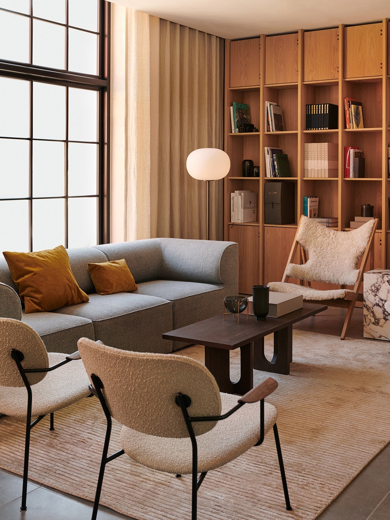Two Co Lounge Chairs with bouclé upholstery in a modern living room setting with a sofa, bookshelf, and large window.