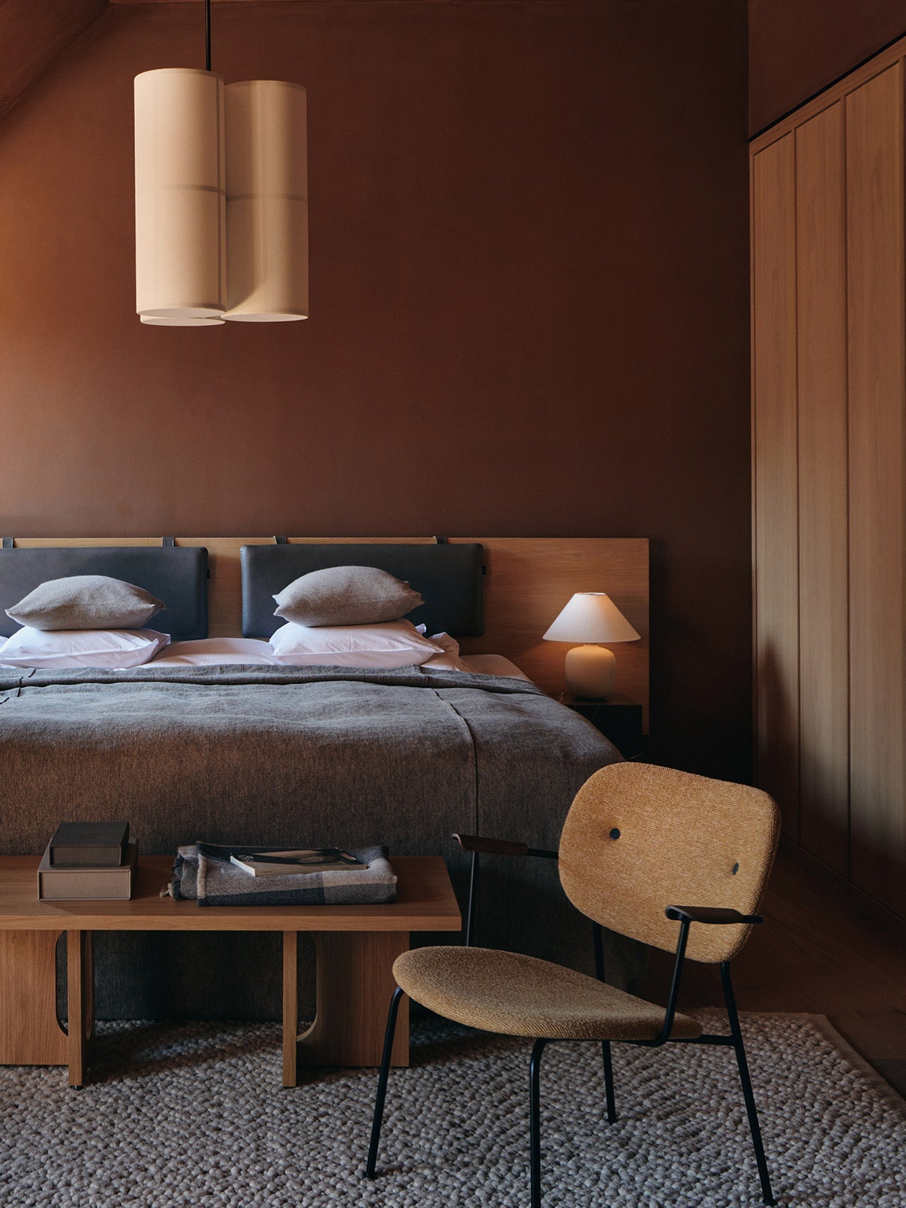 A fully upholstered Co Lounge Chair in a warm, dimly lit bedroom setting with a large bed and dark brown walls.