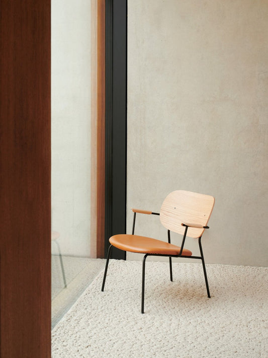 The Co Lounge Chair, with a natural oak backrest, tan leather seat, and black frame, sits on a thick, cream-coloured rug.