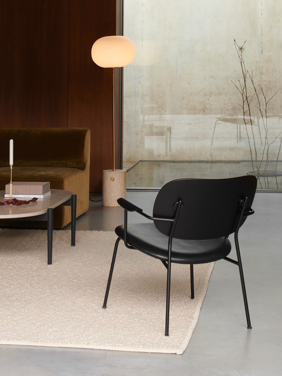 The Co Lounge Chair in black, seen from behind, in a minimalist living room with sofa, coffee table, and glowing floor lamp.