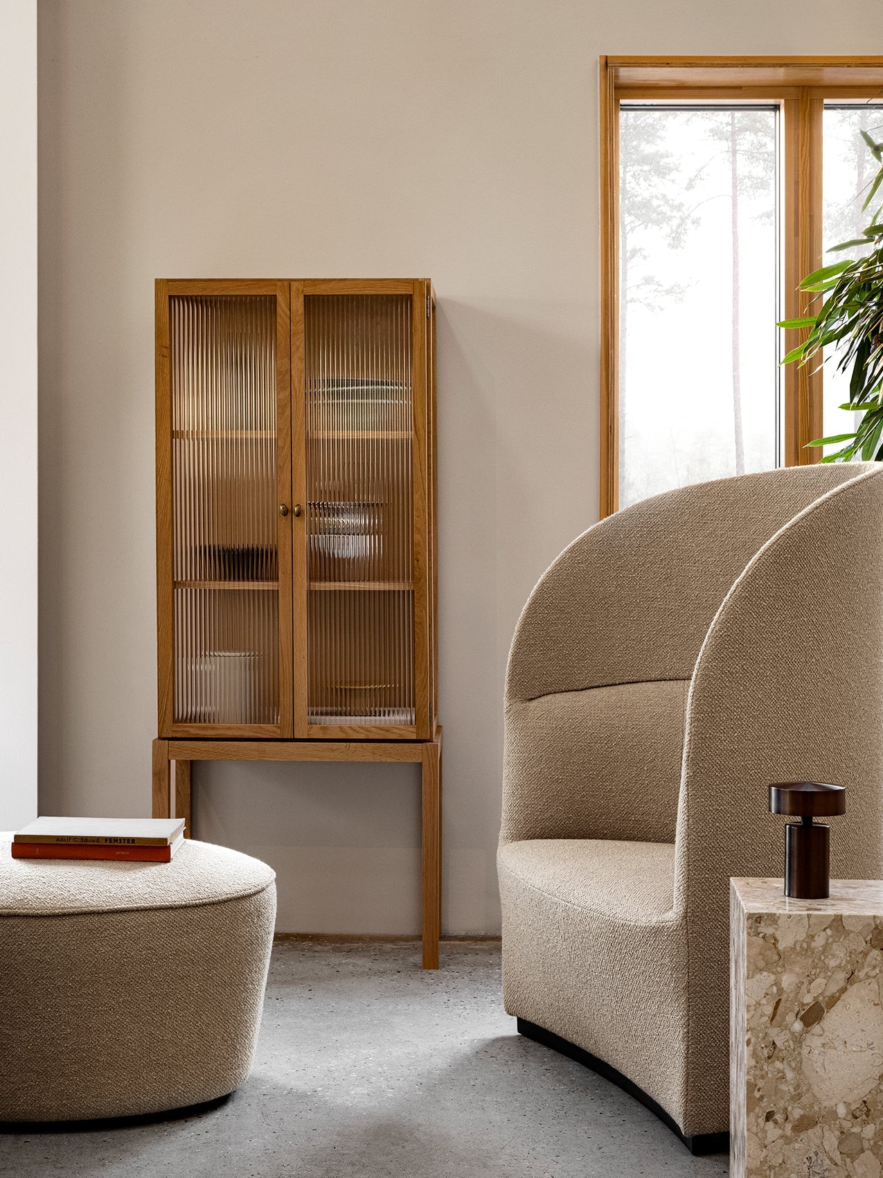 A beige Tearoom Lounge Chair next to a wooden Curiosity Cabinet, with a Cairn Pouf and a Column Table Lamp on a marble Plint.