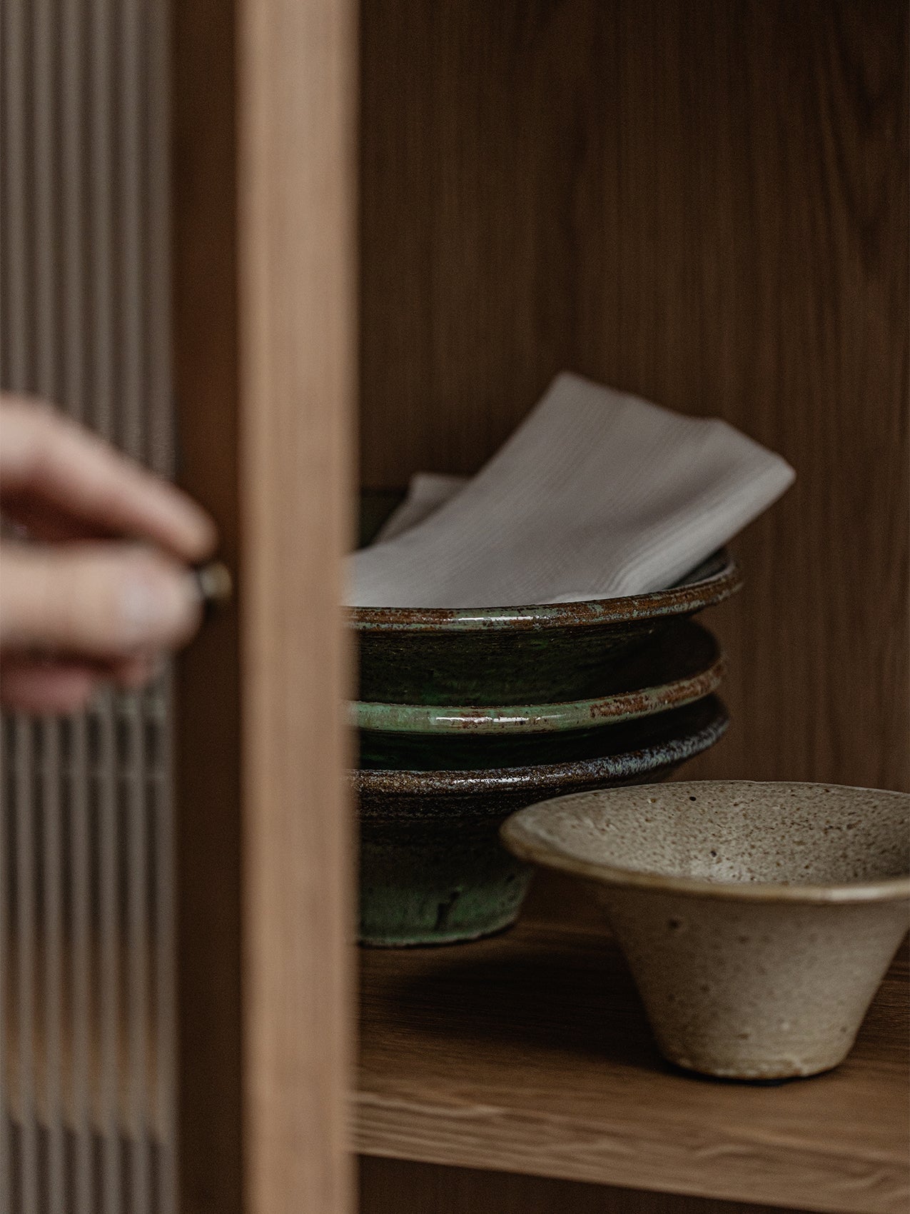 A close-up of a hand opening the oak Curiosity Cabinet, revealing stacked rustic green and beige ceramic bowls on a shelf.