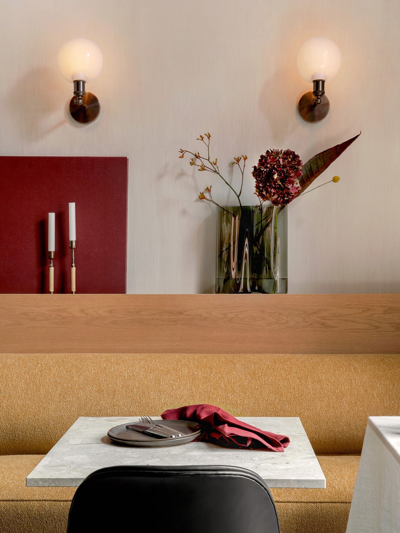 A mustard Eave Dining Sofa at a marble table set with a plate and red napkin. Behind, a ledge holds a vase and candles.