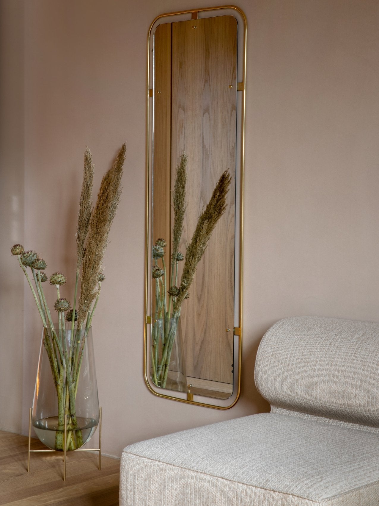 A beige Eave Modular Sofa next to an Echasse Vase with dried flowers. On the wall behind hangs a brass-framed Nimbus Mirror.