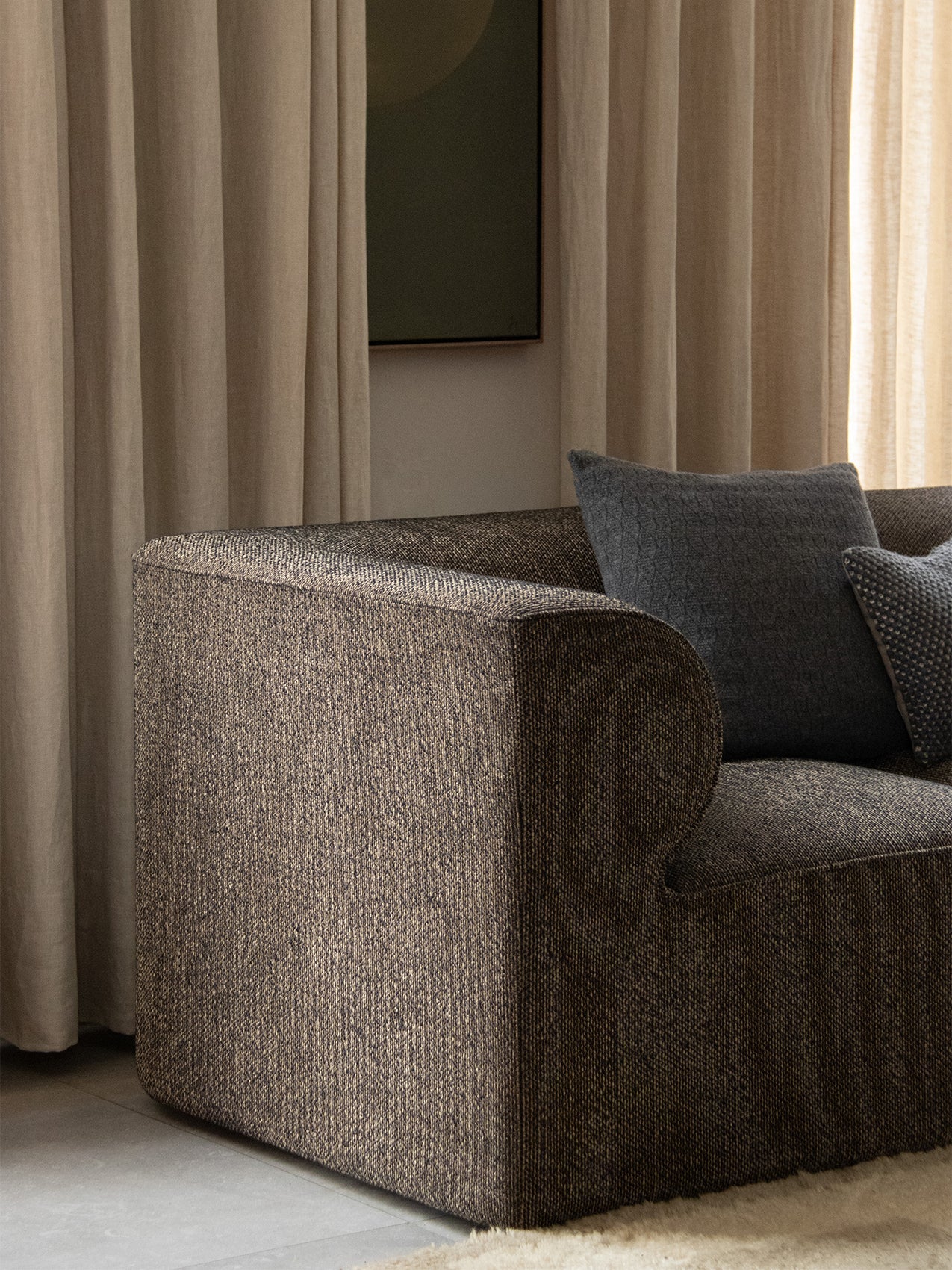 A close-up of the Eave Modular Sofa's textured dark fabric and curved armrest, with two grey cushions against beige curtains.