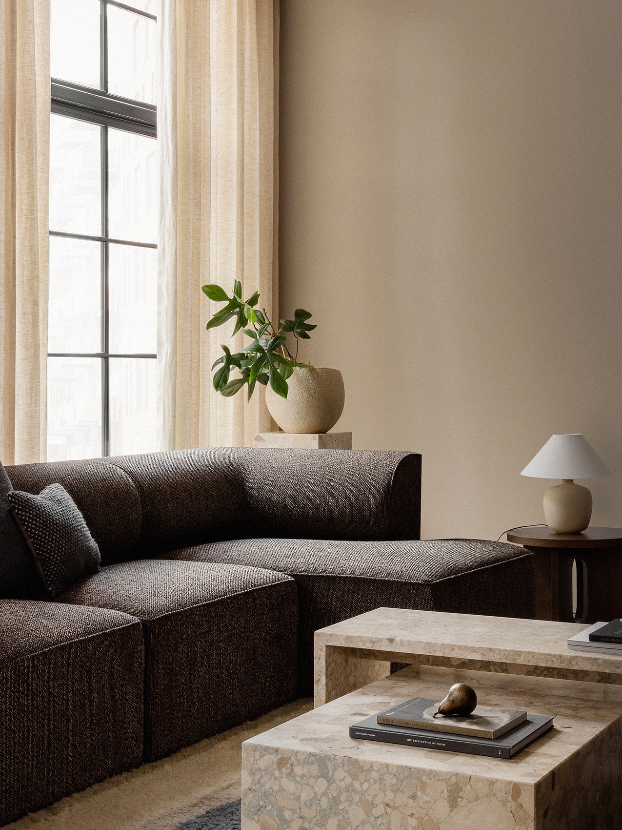 The brown Eave Modular Sofa in a serene living room, styled with stone Plinth coffee tables, a Plantas Planter, and a lamp.